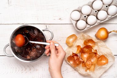 Easter eggs, the process of coloring with natural dye, onion husks in a small saucepan on a white wooden background and a woman's hand holding a colored egg in a spoon.
