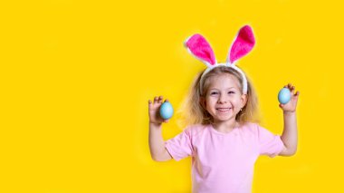 Paskalya tavşanı kulaklı mutlu bir sarışın gülümsüyor ve sarı arka planda Paskalya yumurtalarını tutuyor. Fotokopi alanı, dini bayram konsepti..