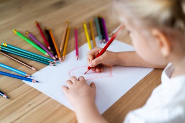 Küçük bir kız masaya renkli kalemlerle kırmızı bir kalp çiziyor..