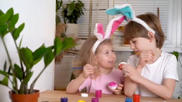 Una niña y un niño con las orejas de liebres de Pascua pintan huevos para las vacaciones con pinceles y pinturas sentados a la mesa en casa — Vídeo de stock