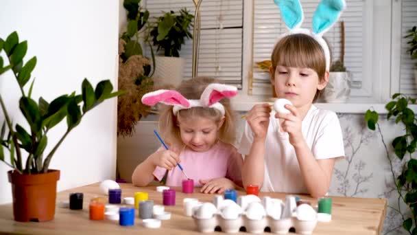 Mała dziewczynka i chłopiec z uszami zające wielkanocne malują jaja na święta pędzlami i farbami siedzącymi przy stole w domu — Wideo stockowe