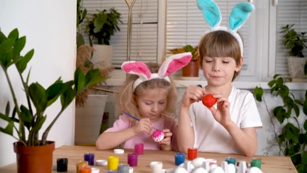 Une petite fille et un garçon aux oreilles de lièvres de Pâques peignent des œufs pour les vacances avec des pinceaux et des peintures assis à la table à la maison — Video