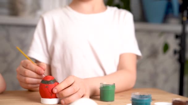Ребенок украшает Пасхальное яйцо красками и кистью, сидя за столом, руки крупным планом — стоковое видео