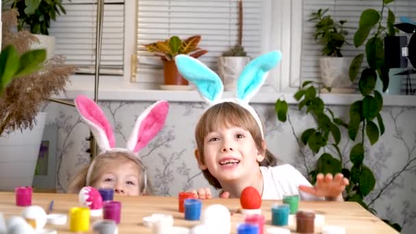 Una niña y un niño con orejas de conejo de Pascua saltan de debajo de la mesa y se ríen, huevos de pollo y pinceles están sobre la mesa. el concepto de fiesta religiosa — Vídeo de stock