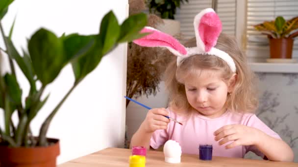 Malá blondýnka zdobí velikonoční vajíčko a usmívá se doma u stolu. koncept náboženského svátku — Stock video