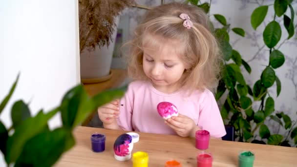 Una niña rubia está decorando un huevo de Pascua y sonriendo, en casa en la mesa. el concepto de fiesta religiosa — Vídeo de stock