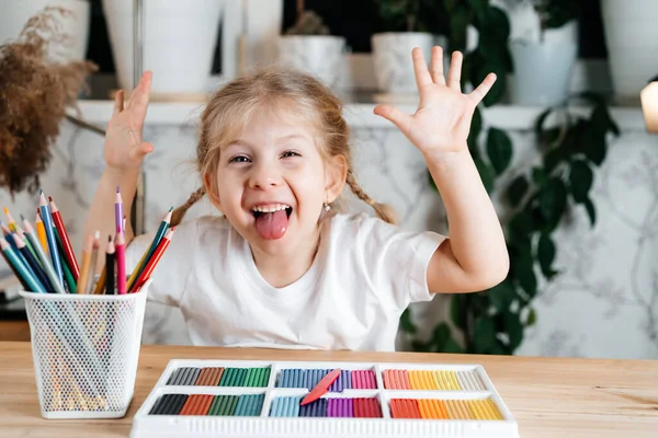 Ένα μικρό ξανθό χαρούμενο κορίτσι γελάει, δείχνει τη γλώσσα της και κάθεται σε ένα τραπέζι με μια φωτεινή πολύχρωμη παλέτα έγχρωμης πλαστελίνης για μόντελινγκ στο σπίτι, χόμπι και την ανάπτυξη των παιδιών. έννοια κατ 'οίκον μάθησης — Φωτογραφία Αρχείου