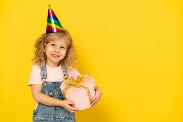 Niña rubia sobre un fondo amarillo sostiene un regalo en una caja redonda rosa con cinta de oro, el concepto de unas vacaciones para niños, espacio para copiar — Foto de Stock