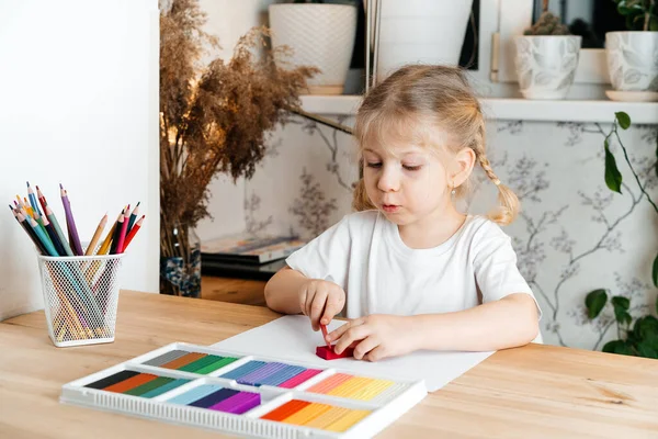 A little blonde girl with a bright colorful palette of colored plasticine for modeling at the table at home, hobbies and child development — Stock Photo, Image