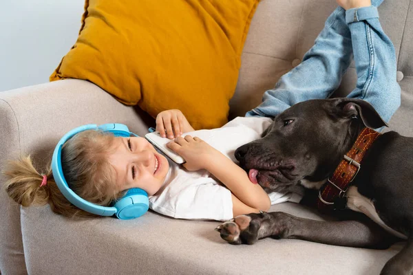 Маленькая Блондинка Слушает Музыку Наушниками Лежа Диване Большой Собакой Улыбаясь — стоковое фото