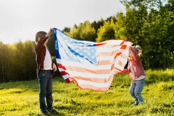 太陽の光に包まれたアメリカ国旗を持つ兄弟姉妹 アメリカ独立記念日の幸せな子供たち — ストック写真