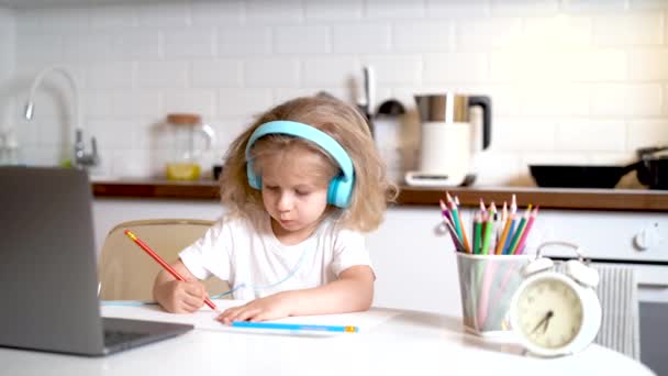 Маленька блондинка в навушниках і перед ноутбуком вдома на кухні малює, вивчаючи концепцію онлайн — стокове відео