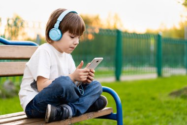 Sokaktaki çocuk, elinde kulaklıkla bankta oturuyor.