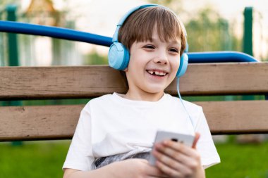 Sokakta oturan mutlu küçük bir çocuk. Kulaklığı, telefonu ve gülümsemesi olan bir bankta oturuyor.