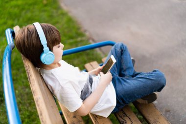 Sokaktaki çocuk, elinde kulaklıkla bankta oturuyor.