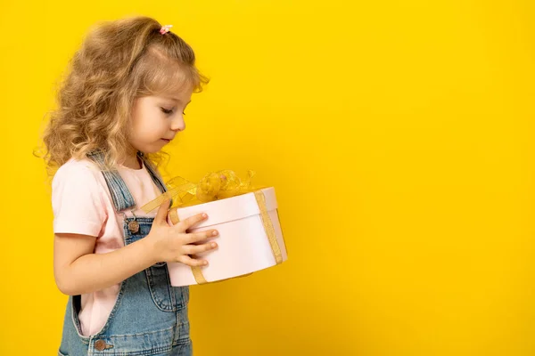 Niña rubia sobre un fondo amarillo sostiene un regalo en una caja redonda rosa con cinta de oro, el concepto de unas vacaciones para niños, espacio para copiar — Foto de Stock