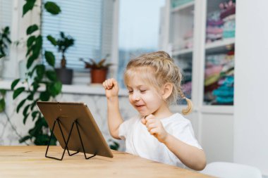 Küçük bir kız elinde tabletle masada oturuyor elleri havada gülümsüyor ve mutlu bir şekilde mutluluğu yaşıyor. Yüksek kalite fotoğraf