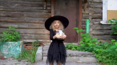Cadılar bayramı için siyah maskeli küçük sarışın bir kız ve eski bir evin arka planında şekerler.
