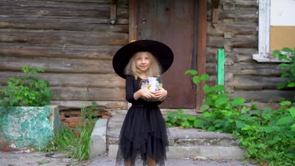 Een klein blond meisje in een zwart gemaskerd kostuum voor Halloween met snoep tegen de achtergrond van een oud huis — Stockvideo