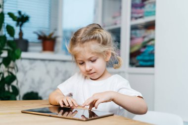 Küçük bir kız elinde tabletle masada oturuyor elleri havada gülümsüyor ve mutlu bir şekilde mutluluğu yaşıyor. Yüksek kalite fotoğraf