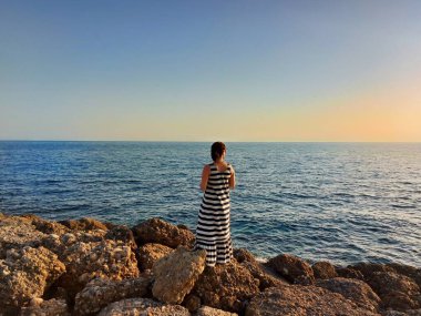 Deniz kenarındaki taşların üzerinde duran bir kadının silueti.