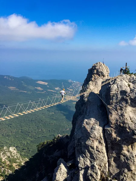 男が緩やかなロープ橋を渡って2つの山の峰の間に広がって歩く ヤルタのアイペトリ山の観光客のための極端なエンターテイメント ストックフォト
