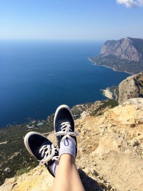 Spor ayakkabı giymiş, yüksek bir dağın tepesinde oturan ve güneşli bir yaz gününde mavi denize ya da okyanusa bakan bir adamın bacakları.