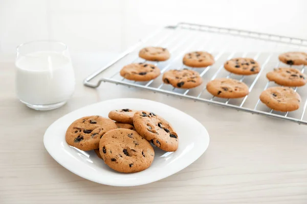 Freshly baked homemade chocolate chip cookies in white plate and on cooling rack with glass of milk on wooden kitchen table. Recipe of cookies with chocolate chips just from oven for lunch treat.