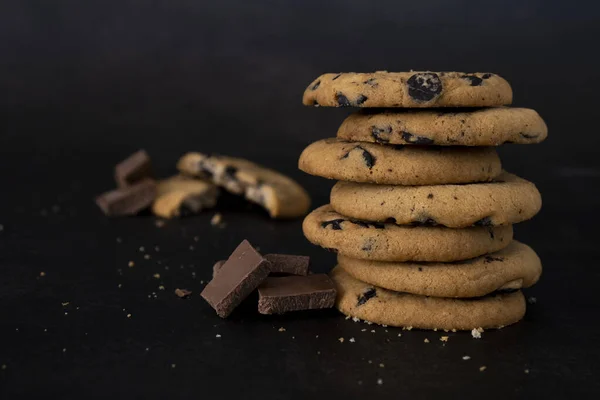 Stack of chocolate chip cookies on black background. Recipe of tasty biscuits with pieces of chocolate. Dessert presentation for restaurant or coffee house. Delicious cookies with chocolate crumbs.