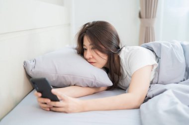 Güzel bir kadın yatak odasındaki yatağın üzerinde cep telefonu görüntülerini kullanıyor.