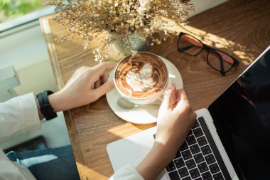 Elinde bir fincan kahve tutan ve tahta masada dizüstü bilgisayarla çalışan bir kadın.