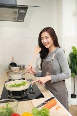 Modern bir mutfakta sebze pişiren mutlu genç bir kadın gülümserken ve elinde tahta bir kaşık tutarken sağlıklı yemek hazırlayan bir fotoğraf.