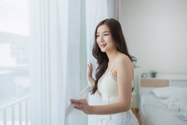 Pencerenin kenarında duran genç kadın, güneşli bir sabah güneşinin tadını çıkarıyor, rahat yatak odasında, huzurlu bir atmosfer ve samimi bir an için katıksız perdeler.