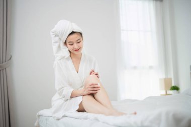 Beyaz bornozlu rahat bir kadın, sağlıklı yaşam ve kendine özen konsepti için doğal ışık ve yumuşak yatak eşliğinde, parlak yatak odasında cilt üstüne losyon sürüyor.