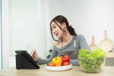 Modern mutfakta dijital tablet kullanan genç bir kadın taze sebzeler ve meyvelerle sağlıklı yemekler hazırlarken, yaşam tarzı, teknoloji, yemek pişirme