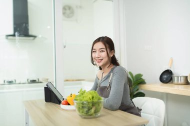 Genç bir kadın gülümserken, modern mutfakta taze sebzeler ve tabletle yemek hazırlıyor.