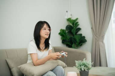 Evde video oyunlarından hoşlanan genç bir kadın, keyifle ve heyecanla kanepede rahatça oturuyor, bitkiler ve dekorlarla dolu modern oturma odası.