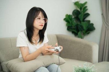 Genç bir kadın kanepede oturmuş oyun kumandasıyla oyun oynuyor, gözlük takıyor ve rahat bir oturma odasında perdeye dikkatlice bakıyor.
