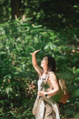 Doğanın tadını çıkaran kadın, yemyeşil ormanda yürürken güneş ışığı yüzünde ve sırt çantasında parlıyor, bir mutluluk ve huzur anı yakalıyor.