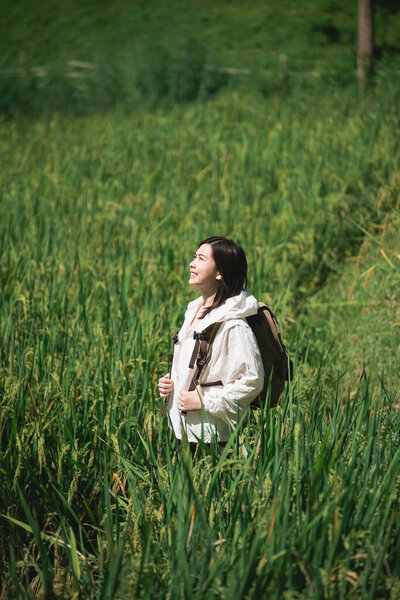 Молодая женщина наслаждается природой, стоя в пышном зеленом поле, улыбаясь и пропитавшись солнечным светом с рюкзаком в солнечный день на открытом воздухе