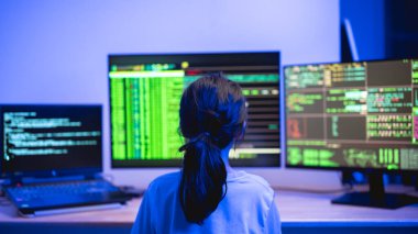 Mavi ışıklandırma efektli loş bir ortamda kod ve veri analizi yapan birden fazla bilgisayar ekranı ile meşgul olan genç kişi