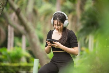 Genç bir kadın yeşil bir bahçede dinlenmeyi seviyor, kulaklık takıyor ve doğanın güzellikleriyle çevrili, müzik dinlemek için akıllı telefonunu kullanıyor..