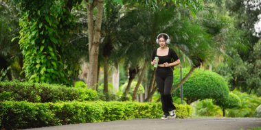 Genç bir kadın yemyeşil bir parkta koşuyor, kulaklık takıyor ve içki içiyor. Bu sahne, doğada aktif ve sağlıklı bir yaşam tarzının özünü yakalar..