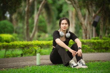 Genç bir kadın canlı bir parkta huzurlu bir anın tadını çıkarıyor. Kulaklıklar ve içeceklerle dolu rahat bir yaşam tarzı sergiliyor..