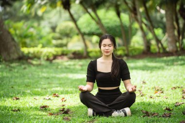 Huzurlu bir parkta oturan genç bir kadın yemyeşil alanda meditasyon yapıyor. Bu görüntü, doğadaki huzuru, sağlığı ve farkındalığın güzelliğini iletir..