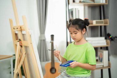 Evde yaratıcı resim aktiviteleri yapan genç bir kız suluboya palet ve sehpa ile sanatsal yetenekler sergiliyor.