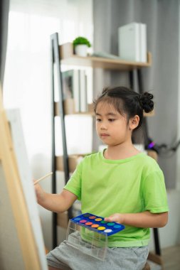 Kapalı bir stüdyoda suluboya resim yapan genç bir kız, odaklanmış ve yaratıcı bir an, doğal ışık ve modern dekorla yakalanmış.