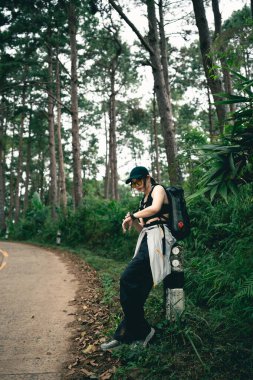 Yeşillik bir ormanda ağaçlarla kaplı bir patikanın yanında duran maceraperest kişi, bir keşif ruhu ve açık hava yaşam tarzı sergiliyor.