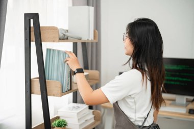 Genç bir kadın modern ofis alanındaki kitaplığı minimalist tasarım ve zarif dekorla düzenliyor, evde verimli bir çalışma ortamının keyfini çıkarıyor.