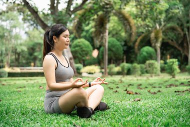 Yeşillik ve rahatlama için ağaçlarla çevrili sakin bir bahçe ortamında meditasyon yapan genç bir kadın.